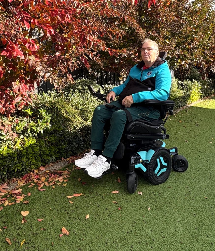 John Duthie sitting in a powered wheelchair beside colourful autumn foliage in a garden.